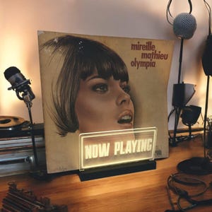 K&ouml;nnte beinhalten: Eine Vintage-Schallplatte mit dem Gesicht einer Frau auf dem Cover. Die Schallplatte tr&auml;gt den Titel "Mireille Mathieu Olympia". Vor der Schallplatte befindet sich ein Neon-Schild mit der Aufschrift "NOW PLAYING".