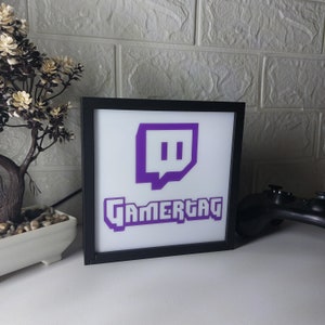 May include: A black framed light-up sign with a purple Twitch logo and the word "GAMERTAG" in purple letters.