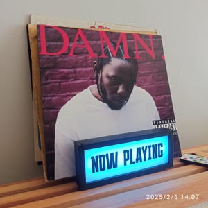 May include: A vinyl record album with the word "DAMN." in red letters. A black and blue "NOW PLAYING" light box sits in front of the album. A remote control is on the right side of the image.