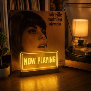 May include: A rectangular acrylic sign with the text NOW PLAYING illuminated in warm yellow light, mounted on a dark wooden base. The sign is positioned in front of a vinyl record cover featuring a portrait of Mireille Mathieu titled Olympia.