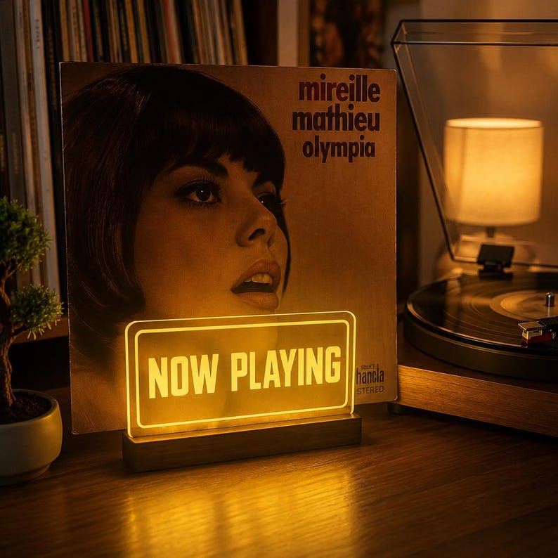 May include: A rectangular acrylic sign with the text NOW PLAYING illuminated in warm yellow light, mounted on a dark wooden base. The sign is positioned in front of a vinyl record cover featuring a portrait of Mireille Mathieu titled Olympia.