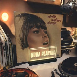 K&ouml;nnte beinhalten: Eine Vintage-Schallplatte mit dem Gesicht einer Frau auf dem Cover. Die Schallplatte tr&auml;gt den Titel "Mireille Mathieu Olympia". Vor der Schallplatte befindet sich ein Leuchtreklameschild mit der Aufschrift "NOW PLAYING".