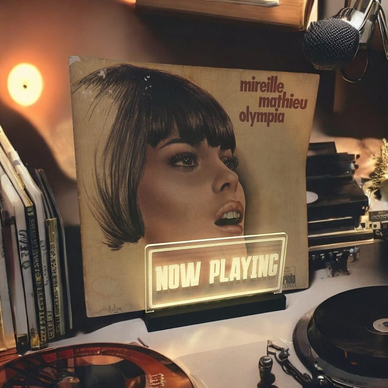 K&ouml;nnte beinhalten: Eine Vintage-Schallplatte mit dem Gesicht einer Frau auf dem Cover. Die Schallplatte tr&auml;gt den Titel "Mireille Mathieu Olympia". Vor der Schallplatte befindet sich ein Leuchtreklameschild mit der Aufschrift "NOW PLAYING".