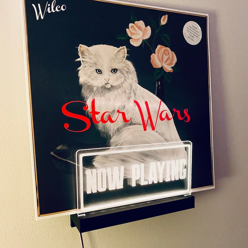 K&ouml;nnte beinhalten: Eine wei&szlig;e Katze mit schwarzer Nase und rosa Ohren sitzt auf einer schwarzen Oberfl&auml;che. Die Katze schaut in die Kamera. Der Hintergrund ist ein Gem&auml;lde mit Rosen und dem Text "Wilco Star Wars". Ein wei&szlig;es Schild mit dem Text "NOW PLAYING" befindet sich vor der Katze.