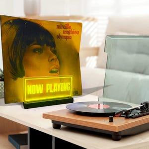 K&ouml;nnte beinhalten: Ein gelbes Neon-Schild mit der Aufschrift "NOW PLAYING" steht vor einer Vintage-Schallplatte mit einer Frau auf dem Cover. Die Schallplatte tr&auml;gt den Titel "Mireille Mathieu Olympia".