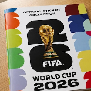 Op de afbeelding: Een album van de officiële stickercollectie voor het Wereldkampioenschap 2026. De cover toont de FIFA World Cup-trofee, het FIFA-logo en de tekst "WORLD CUP 2026". De cover heeft een witte achtergrond met kleurrijke abstracte vormen.