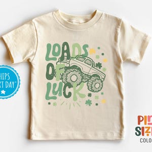 May include: Cream-colored toddler t-shirt with a green monster truck graphic and the words "Loads of Luck." The design includes shamrocks and stars. The shirt is made of soft material.