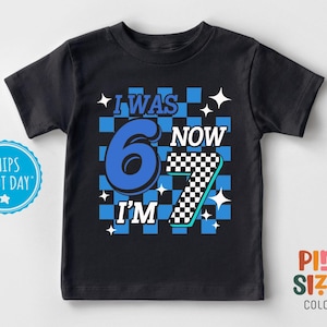 May include: Black t-shirt with the text "I WAS 6 NOW I'M 7" in blue, white, and black. The number 6 is blue, and the number 7 is black and white checkered. The background has a blue checkered pattern.
