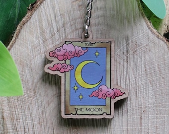 Könnte beinhalten: Ein Holzschlüsselanhänger mit einem Tarotkarten-Design. Die Karte zeigt einen Halbmond, Sterne und rosa und blaue Wolken. Der Schlüsselanhänger ist an einer silbernen Kette und einem Ring befestigt. Der Text "THE MOON" ist sichtbar.