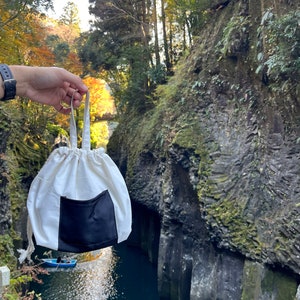 May include: A white drawstring bag with a black pocket. The bag is hanging from a person's hand in front of a scenic view of a river and cliffs.