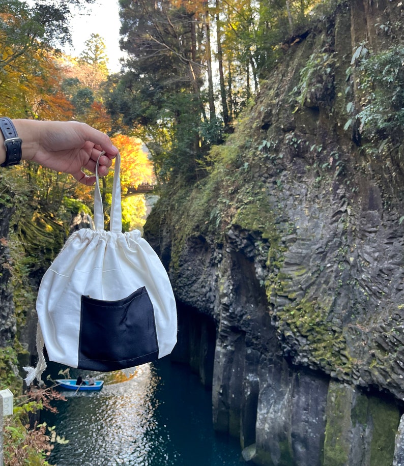 May include: A white drawstring bag with a black pocket. The bag is hanging from a person's hand in front of a scenic view of a river and cliffs.