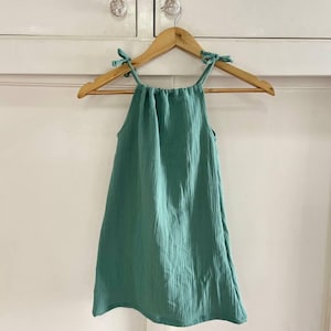 May include: A teal sleeveless dress with a gathered neckline and tie straps. The dress is hanging on a wooden hanger.