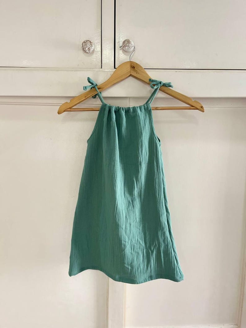 May include: A teal sleeveless dress with a gathered neckline and tie straps. The dress is hanging on a wooden hanger.