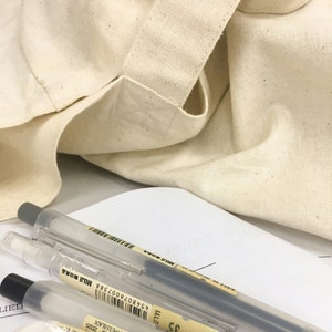 以下が含まれることがあります： 白い表面に置かれた、グレーと黒のキャップが付いた3本の透明なプラスチックペン。背景にはベージュのキャンバス地のトートバッグ。ペンには「MUJI」のブランド名が印刷されています。