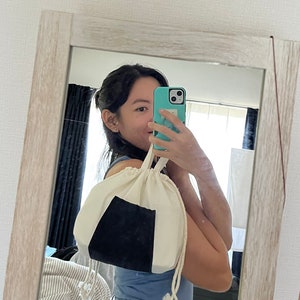 May include: A person is holding a white drawstring bag with a black triangle on it. The bag is made of a soft, natural fabric. The person is wearing blue jeans and a blue shirt.