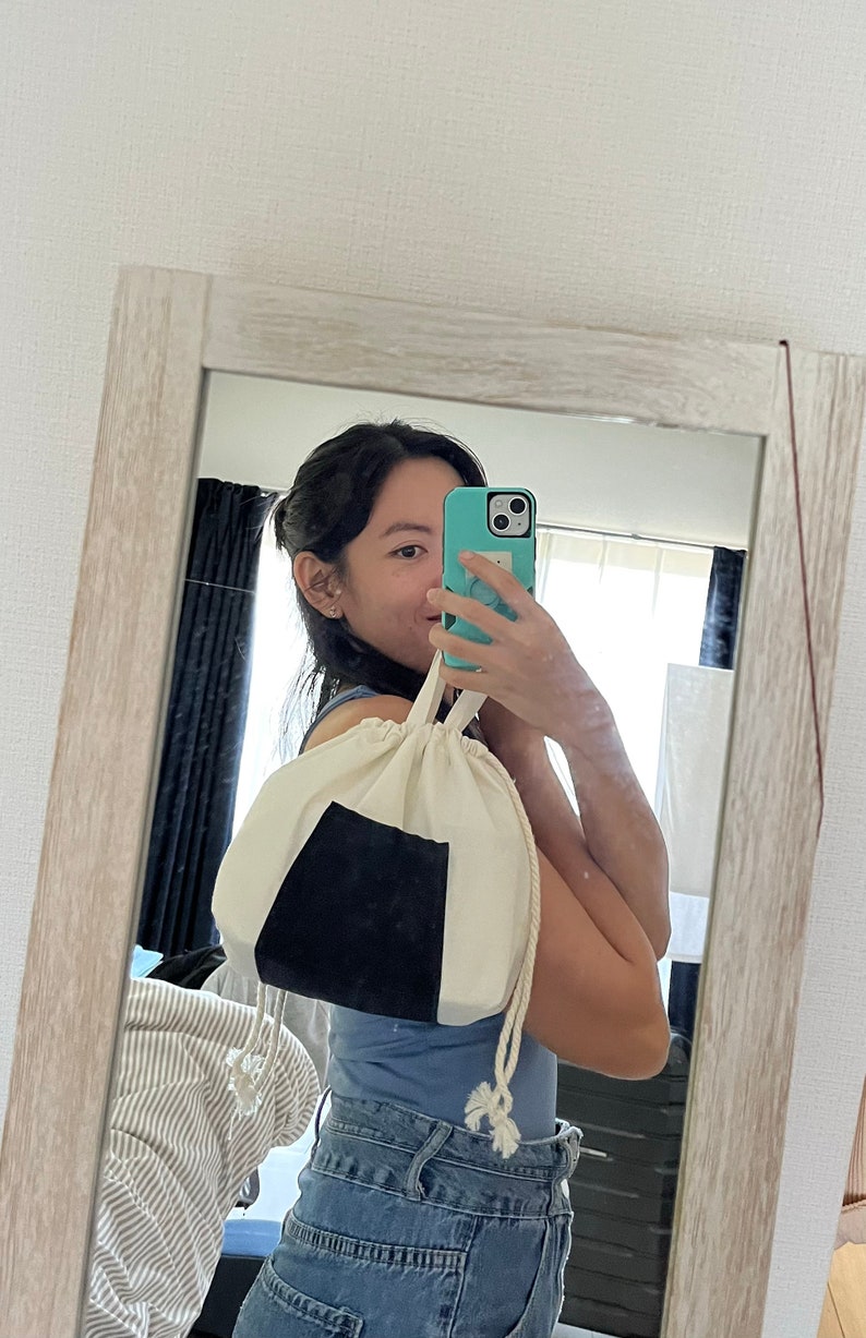 May include: A person is holding a white drawstring bag with a black triangle on it. The bag is made of a soft, natural fabric. The person is wearing blue jeans and a blue shirt.