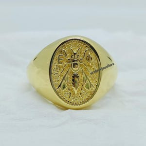 May include: A gold signet ring with an oval shaped top featuring an engraved design of a bee with wings outstretched. The bee is surrounded by a decorative border.