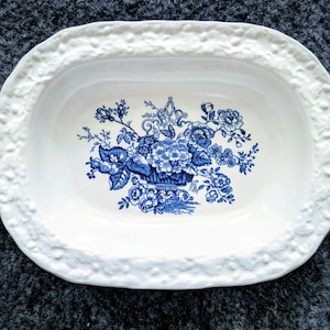 May include: White oval serving dish with a blue floral pattern. The central design shows a basket of flowers and other floral motifs. The rim of the dish has a textured, raised border.