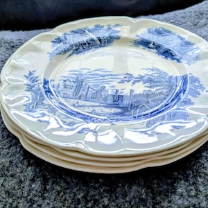 May include: A stack of white ceramic plates with a blue and white transferware design. The plates have a scalloped edge and a detailed landscape scene with buildings and trees.