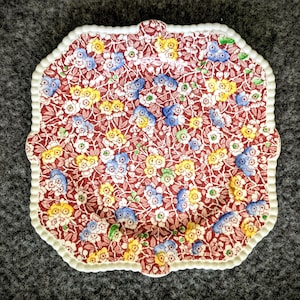 May include: A square plate with a scalloped edge, decorated with a floral pattern. The pattern includes red, blue, yellow, and white flowers. The edge of the plate is white. The plate is likely made of ceramic or porcelain.