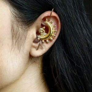 May include: Gold-toned ear cuff featuring a crescent moon design with sun-like embellishments and a small bird figure. The ear cuff wraps around the ear and is secured with a thin wire. The jewelry is worn on the ear.