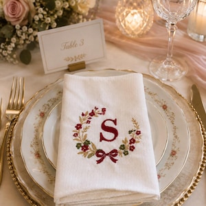 Könnte beinhalten: Eine weiße Stoffserviette mit einer Stickerei aus einem Blumenkranz in rotem und grünem Garn, in dessen Mitte ein großer roter Buchstabe S und unten eine kleine rote Schleife zu sehen sind. Die Serviette ist gefaltet und liegt auf einem weißen Teller.