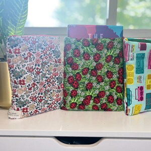 Könnte beinhalten: Drei Buchhüllen aus Stoff mit einzigartigen Mustern. Eine Hülle zeigt ein Blumenmuster mit roten, rosa und grünen Blumen auf weißem Grund. Eine andere hat ein Marienkäfermuster auf grünem Grund. Die dritte hat ein Retro-Boombox-Design.