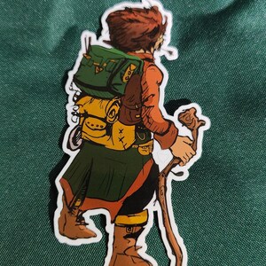 May include: A die-cut sticker featuring a cartoon illustration of a traveler with brown hair, wearing an orange shirt, a dark green skirt, and brown boots. The figure carries a large green and yellow backpack and holds a wooden walking staff.