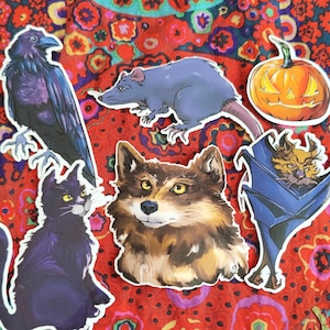Könnte beinhalten: Eine Sammlung von Aufklebern mit verschiedenen Kreaturen. Enthält eine schwarze Katze, einen Raben, einen Wolf, eine Ratte, einen Kürbis und eine Fledermaus. Die Aufkleber sind vor einem lebendigen, floral gemusterten Hintergrund angeordnet.
