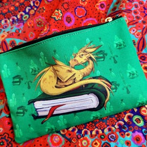 May include: A green zippered pouch featuring a golden dragon resting on a book with a red bookmark. The pouch has a black zipper and a repeating pattern of swords and other shapes. The background is a colorful floral pattern.