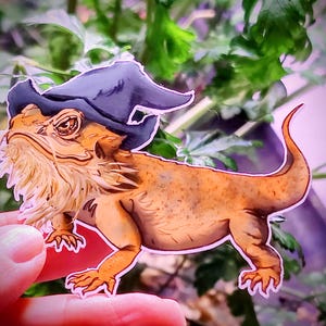 May include: A cartoon illustration of a bearded dragon wearing a purple and black wizard hat. The dragon is orange and brown with a long tail and is standing on its hind legs.