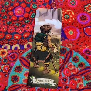 May include: A bookmark featuring an illustrated adventurer with a backpack and walking stick, set against a mountain backdrop. The character is wearing a brown jacket and green backpack. The word "Adventure" is written on a banner at the bottom.