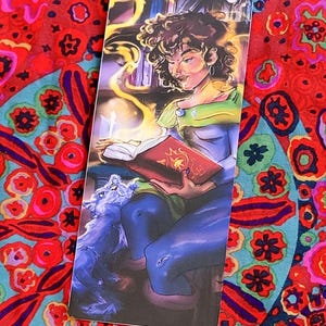 May include: A rectangular bookmark featuring an illustration of a person reading a book with a wolf. The person has curly brown hair and is wearing a green and blue outfit. The book is red, and yellow light emanates from it. The background is a library.