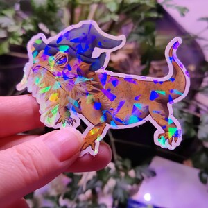 May include: A holographic sticker of a bearded dragon wearing a blue wizard hat. The sticker has a glittery, iridescent finish.