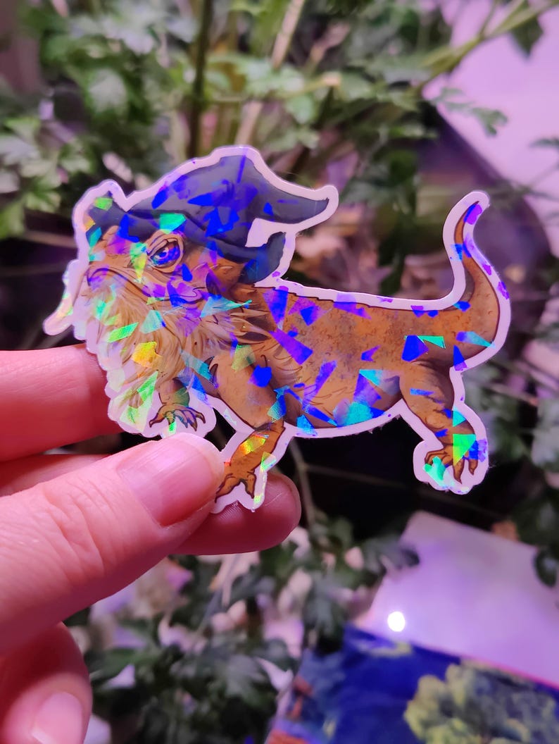 May include: A holographic sticker of a bearded dragon wearing a blue wizard hat. The sticker has a glittery, iridescent finish.