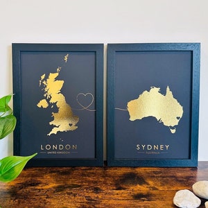 May include: Two black framed prints with gold foil maps of London, United Kingdom and Sydney, Australia. The maps are connected by a gold foil line with a heart at the end.