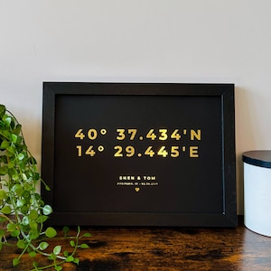 May include: A black framed print with gold lettering. The print features the coordinates 40° 37.434' N 14° 29.445' E and the text "Shen & Tom Positano, IT - 03.08.2021".
