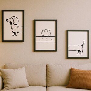 May include: Three framed art prints with black frames hanging above a beige sofa. The prints feature minimalist black and white illustrations of a dachshund dog and a bowl of plants. The art adds a touch of whimsy to the room.
