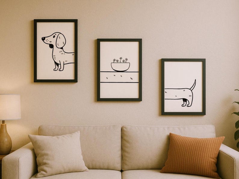 May include: Three framed art prints with black frames hanging above a beige sofa. The prints feature minimalist black and white illustrations of a dachshund dog and a bowl of plants. The art adds a touch of whimsy to the room.