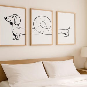 May include: Three framed art prints with a minimalist dachshund design. The prints, in light wood frames, show the dog in various poses, using black line art on a white background. The artwork is displayed above a bed, adding a touch of modern art.