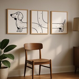 May include: Three framed art prints with black and white dachshund illustrations. The prints are in light wood frames, displayed on a neutral wall. A wooden chair and a potted plant are in the foreground, with a lamp and cabinet in the background.