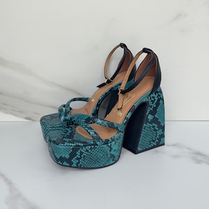 May include: Teal platform sandals with a snake print design and black heels. The sandals have a thick ankle strap and a criss-cross strap across the toes.