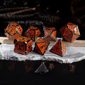 Puede incluir: Conjunto de siete dados de metal naranja y negro con diseños intrincados. Los dados están numerados del 4 al 20.