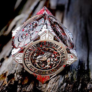 May include: A multi-sided, silver-colored die with intricate designs and red accents. The die features circular faces with numbers and detailed illustrations, possibly for a game or decorative item. The die is resting on a textured wooden surface.