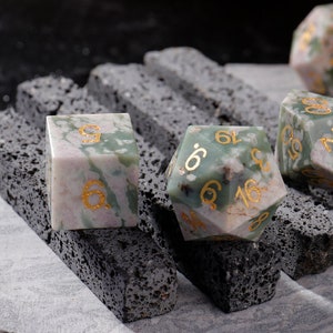 May include: A set of six-sided dice made of green and white speckled stone with gold numbers. The dice are sitting on a black, textured surface.