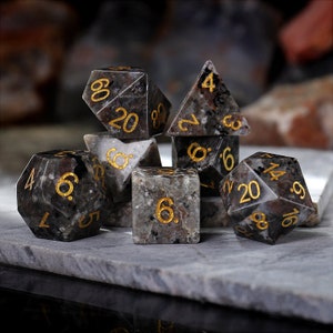 May include: A set of seven black and grey gemstone dice with gold numbers. The dice are in various shapes, including a pyramid, a cube, and a dodecahedron.