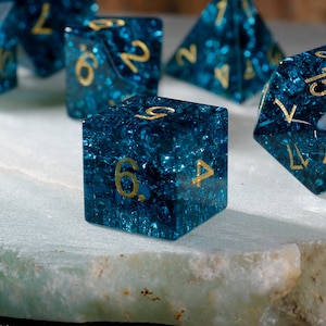 Peut inclure: Un ensemble de dés bleus et dorés avec une finition scintillante. Les dés sont numérotés de 1 à 6 et sont faits d'un matériau transparent avec une teinte bleue. Les dés sont posés sur une surface blanche.