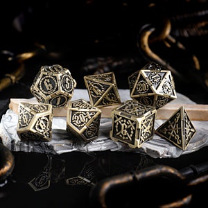 Puede incluir: Un conjunto de siete dados de metal dorado con marcas negras. Los dados son de diferentes formas y tamaños, y cada uno tiene un diseño diferente. Los dados están sobre una superficie gris.