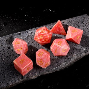 Puede incluir: Conjunto de siete dados rosas y rojos con números dorados. Los dados están hechos de piedra natural y tienen un patrón único de mármol.