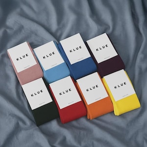 May include: Eight pairs of folded socks in various solid colors including pink, light blue, dark blue, dark purple, black, red, orange, and yellow. Each pair features a white paper label with the black text KLUE printed on the front.
